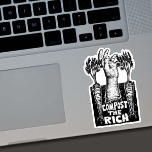 May include: Black and white sticker featuring a hand emerging from the ground between two carrots. The text "COMPOST THE RICH" is printed below the hand. The sticker is placed on a laptop.