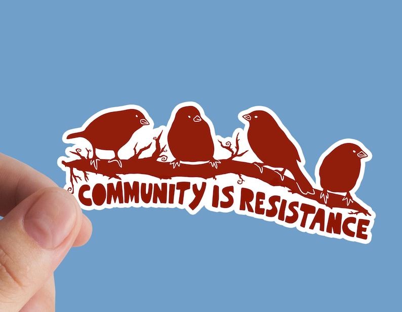 May include: A sticker featuring four stylized brown birds perched on a brown branch. The text "COMMUNITY IS RESISTANCE" is printed in white below the birds. The sticker is held against a light blue background.