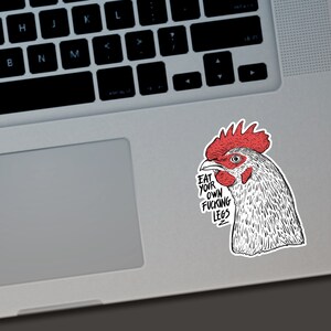 May include: A sticker featuring a detailed illustration of a rooster's head with a red comb and wattle. The sticker has the text "EAT YOUR OWN FUCKING LEGS" in a bold, black font. The sticker is placed on a laptop.