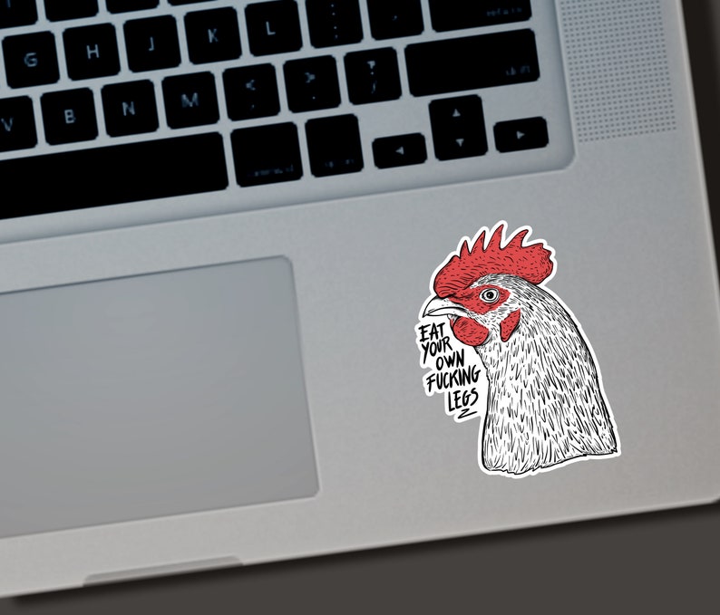 May include: A sticker featuring a detailed illustration of a rooster's head with a red comb and wattle. The sticker has the text "EAT YOUR OWN FUCKING LEGS" in a bold, black font. The sticker is placed on a laptop.