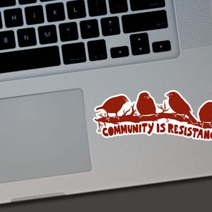 May include: A sticker featuring four stylized birds perched on a branch, in a reddish-brown color. The text "COMMUNITY IS RESISTANCE" is below the birds. The sticker is placed on a laptop.