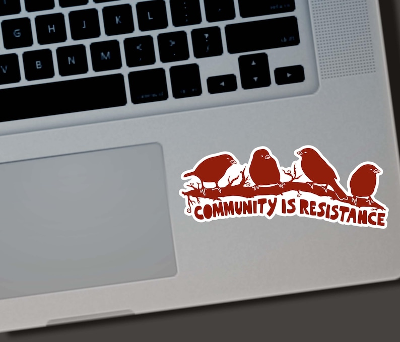 May include: A sticker featuring four stylized birds perched on a branch, in a reddish-brown color. The text "COMMUNITY IS RESISTANCE" is below the birds. The sticker is placed on a laptop.