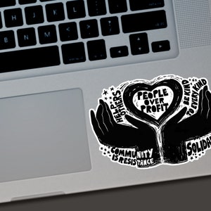 May include: Black and white sticker with the text "People Over Profit" inside a heart shape, held by two hands. Additional text includes "Help Others", "Community is Resistance", "Be Kind to Every Kind", and "Solidarity". The sticker is on a laptop.