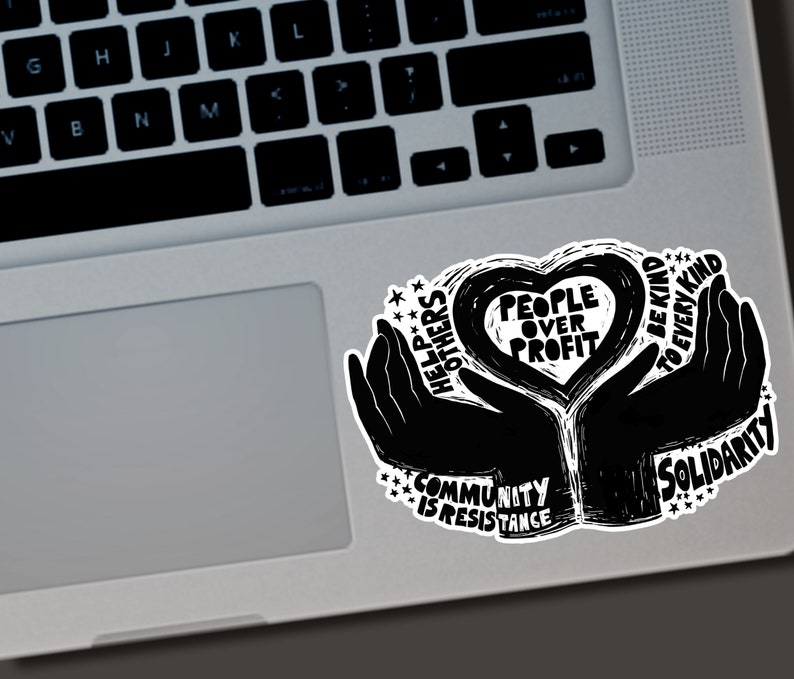 May include: Black and white sticker with the text "People Over Profit" inside a heart shape, held by two hands. Additional text includes "Help Others", "Community is Resistance", "Be Kind to Every Kind", and "Solidarity". The sticker is on a laptop.