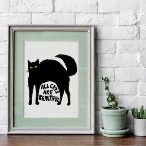 May include: Framed art print featuring a black cat silhouette with yellow eyes. The cat's arched back forms the shape of the text "ALL CATS ARE BEAUTIFUL" in white. The artwork is framed in a light green and gray frame, set against a white brick wall.