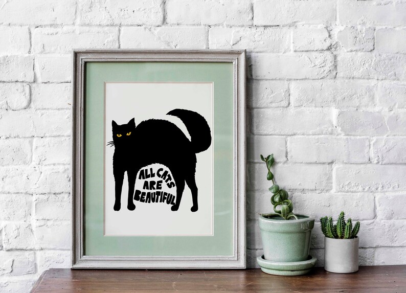 May include: Framed art print featuring a black cat silhouette with yellow eyes. The cat's arched back forms the shape of the text "ALL CATS ARE BEAUTIFUL" in white. The artwork is framed in a light green and gray frame, set against a white brick wall.
