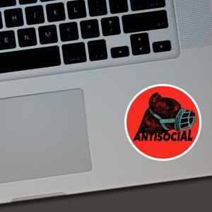 May include: A round sticker with a red background featuring a black and teal illustration of a dog wearing a muzzle. The word "ANTISOCIAL" is printed in white below the dog illustration.