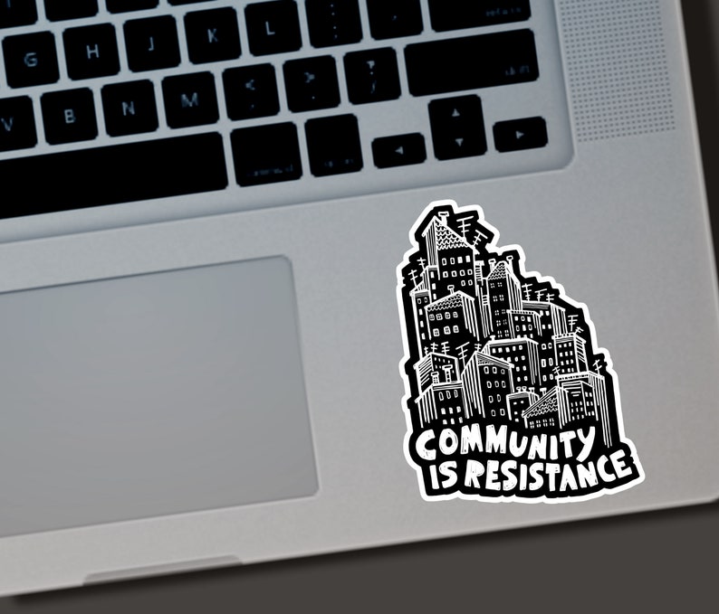 May include: A black and white sticker featuring a cityscape illustration. The sticker has the words "COMMUNITY IS RESISTANCE" in bold, white text. The sticker is placed on a laptop.