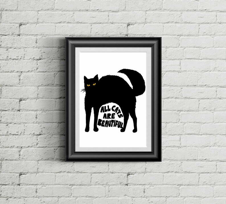 May include: Black framed art print featuring a black cat silhouette with yellow eyes. The text "ALL CATS ARE BEAUTIFUL" is printed in white within the cat's body. The artwork is displayed against a white background and a brick wall.