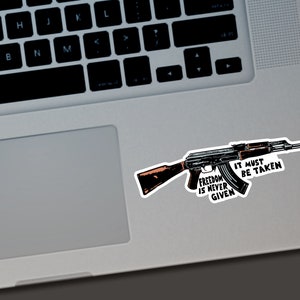 Puede incluir: Una pegatina con una ilustraci&oacute;n detallada de un rifle AK-47. La pegatina tiene el texto "Freedom is Never Given, It Must Be Taken" en una fuente audaz y estilizada. El rifle est&aacute; representado en tonos de negro, marr&oacute;n y gris.
