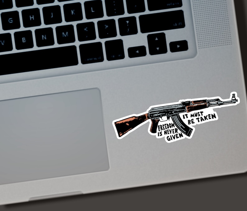 Puede incluir: Una pegatina con una ilustraci&oacute;n detallada de un rifle AK-47. La pegatina tiene el texto "Freedom is Never Given, It Must Be Taken" en una fuente audaz y estilizada. El rifle est&aacute; representado en tonos de negro, marr&oacute;n y gris.