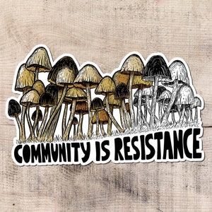 May include: A sticker featuring a cluster of stylized mushrooms in shades of brown, tan, and black. The phrase "COMMUNITY IS RESISTANCE" is written in bold, black letters below the mushrooms. The sticker is on a light wood grain background.