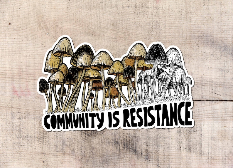 May include: A sticker featuring a cluster of stylized mushrooms in shades of brown, tan, and black. The phrase "COMMUNITY IS RESISTANCE" is written in bold, black letters below the mushrooms. The sticker is on a light wood grain background.