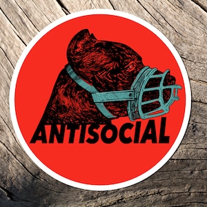 May include: A round sticker with a red background featuring a detailed illustration of a dog's head wearing a muzzle. The word "ANTISOCIAL" is printed in white below the dog.
