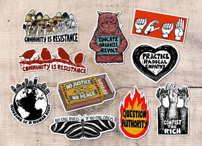 May include: A collection of stickers with various designs and text. Designs include mushrooms, birds on a branch, a cat holding a sign, hands spelling out words, a heart, a globe, a matchbox, a butterfly, flames, and a hand with carrots. The stickers have phrases like "Community is Resistance" and "Question Authority."