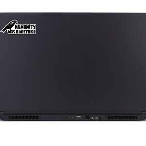 May include: Black laptop with a sticker featuring a black raven and the text "HUMANITY WAS A MISTAKE" in a white rectangular box. The laptop has a sleek, matte finish.