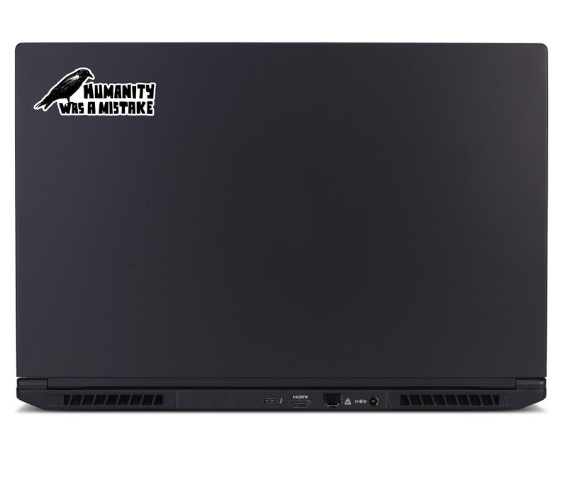 May include: Black laptop with a sticker featuring a black raven and the text "HUMANITY WAS A MISTAKE" in a white rectangular box. The laptop has a sleek, matte finish.