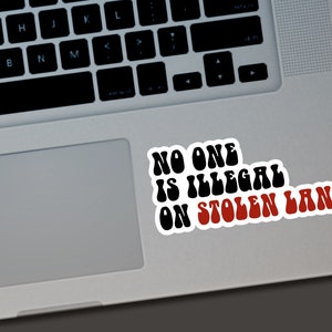 Puede incluir: Una pegatina blanca con el texto "NO ONE IS ILLEGAL ON STOLEN LAND" en negro y rojo, colocada en un port&aacute;til. El texto est&aacute; en una fuente retro en negrita.