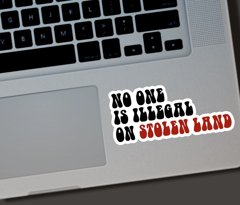Puede incluir: Una pegatina blanca con el texto "NO ONE IS ILLEGAL ON STOLEN LAND" en negro y rojo, colocada en un port&aacute;til. El texto est&aacute; en una fuente retro en negrita.