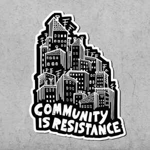 May include: A black and white sticker featuring a stylized cityscape with the text "COMMUNITY IS RESISTANCE" in bold, white letters. The illustration depicts buildings with antennas and various architectural details, creating a graphic design.