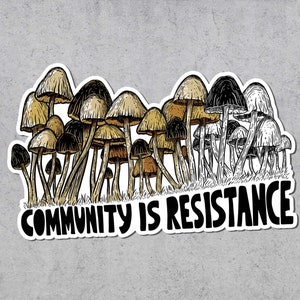 May include: A sticker featuring a cluster of stylized mushrooms in shades of brown, tan, and black. The text "COMMUNITY IS RESISTANCE" is written in bold, black letters below the mushrooms. The sticker has a white border.
