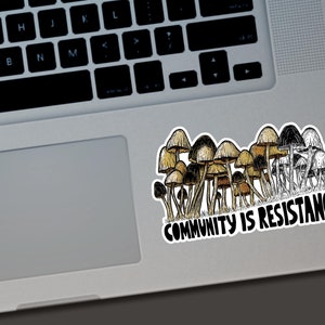 May include: A sticker featuring a cluster of mushrooms in shades of brown, tan, and black, with the text "COMMUNITY IS RESISTANCE" in bold, black letters. The sticker is placed on a laptop.