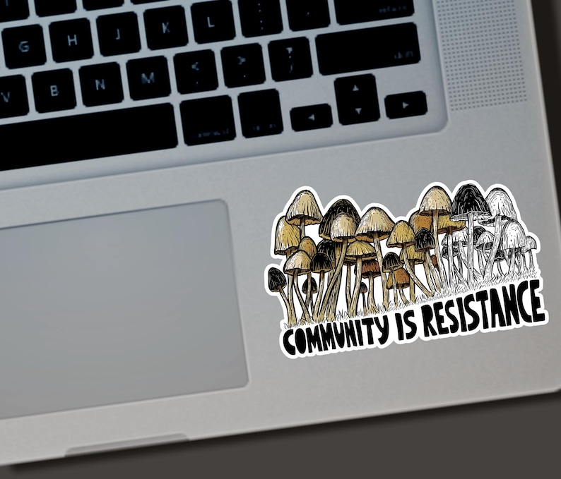 May include: A sticker featuring a cluster of mushrooms in shades of brown, tan, and black, with the text "COMMUNITY IS RESISTANCE" in bold, black letters. The sticker is placed on a laptop.