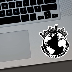 May include: A black and white sticker featuring a globe with a ring of houses around the top. The text "HOUSING IS A HUMAN RIGHT" is printed in a circular pattern around the globe. The sticker is placed on a laptop.