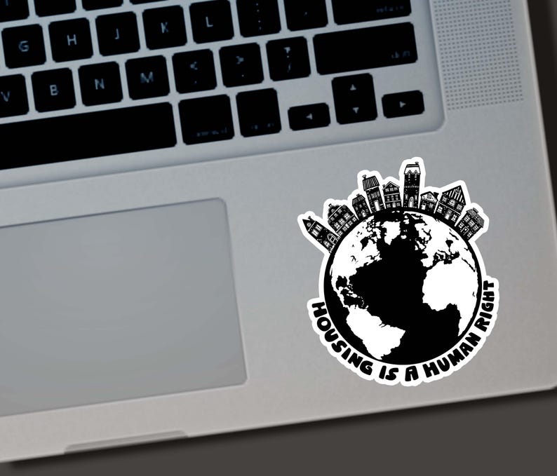 May include: A black and white sticker featuring a globe with a ring of houses around the top. The text "HOUSING IS A HUMAN RIGHT" is printed in a circular pattern around the globe. The sticker is placed on a laptop.