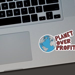 May include: A white sticker with a blue and black globe illustration and the text "PLANET OVER PROFIT" in red. The sticker is placed on a silver laptop.