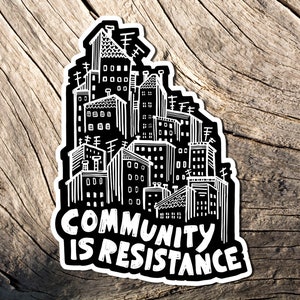 May include: A black and white sticker featuring a cityscape design. The sticker has a white border and the words "COMMUNITY IS RESISTANCE" in bold, white letters. The sticker is placed on a weathered, wooden surface.
