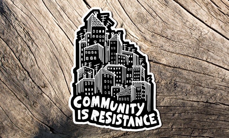 May include: A black and white sticker featuring a cityscape design. The sticker has a white border and the words "COMMUNITY IS RESISTANCE" in bold, white letters. The sticker is placed on a weathered, wooden surface.