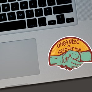 Puede incluir: Una pegatina con el texto "ORGANIZE A REVOLUTION" en un dise&ntilde;o de semic&iacute;rculo. La pegatina presenta un gr&aacute;fico de apret&oacute;n de manos en verde azulado y rojo, sobre un fondo amarillo y naranja. La pegatina est&aacute; colocada en un port&aacute;til.