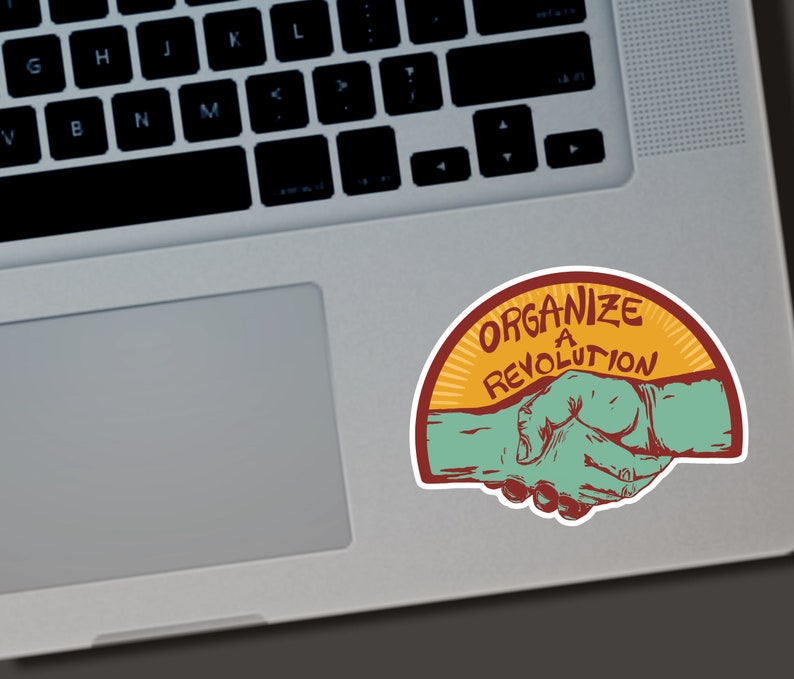 Puede incluir: Una pegatina con el texto "ORGANIZE A REVOLUTION" en un dise&ntilde;o de semic&iacute;rculo. La pegatina presenta un gr&aacute;fico de apret&oacute;n de manos en verde azulado y rojo, sobre un fondo amarillo y naranja. La pegatina est&aacute; colocada en un port&aacute;til.