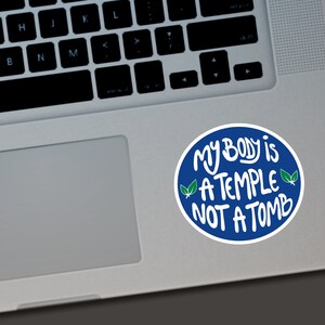 May include: A round sticker with a blue border and white text that reads "MY BODY is A TEMPLE NOT A TOMB." The sticker also features two green leaf illustrations. The sticker is on a laptop.
