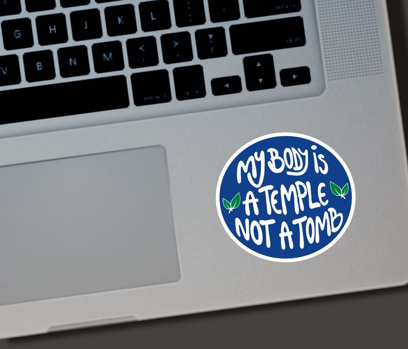 May include: A round sticker with a blue border and white text that reads "MY BODY is A TEMPLE NOT A TOMB." The sticker also features two green leaf illustrations. The sticker is on a laptop.