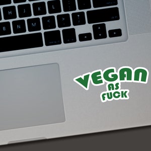 May include: A white sticker with the words "VEGAN AS FUCK" in green, placed on a laptop. The text is in a bold, rounded font with a white outline, and the sticker is slightly angled.