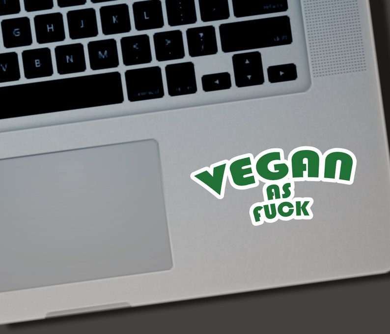 May include: A white sticker with the words "VEGAN AS FUCK" in green, placed on a laptop. The text is in a bold, rounded font with a white outline, and the sticker is slightly angled.