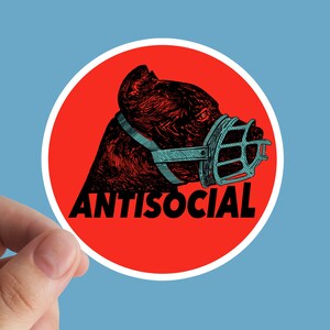 May include: A round sticker featuring a dog's head wearing a muzzle, set against a red circle with a white border. The word "ANTISOCIAL" is printed in bold black letters below the dog's head. The background is light blue.