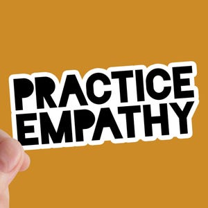 May include: A white sticker with the words "PRACTICE EMPATHY" in bold black letters. The sticker is held against a solid orange background. The sticker has a white border.