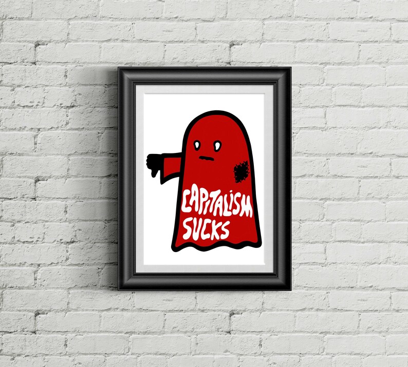 May include: Framed art print featuring a red ghost with a thumbs-down gesture. The ghost has the words "CAPITALISM SUCKS" in white text. The artwork is displayed in a black frame against a white brick wall.