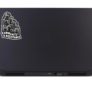 May include: Black laptop with a sticker featuring a cityscape design in white and black. The sticker reads "COMMUNITY IS RESISTANCE" in bold, capital letters. The laptop is closed, showing its back, with a sleek, matte finish.
