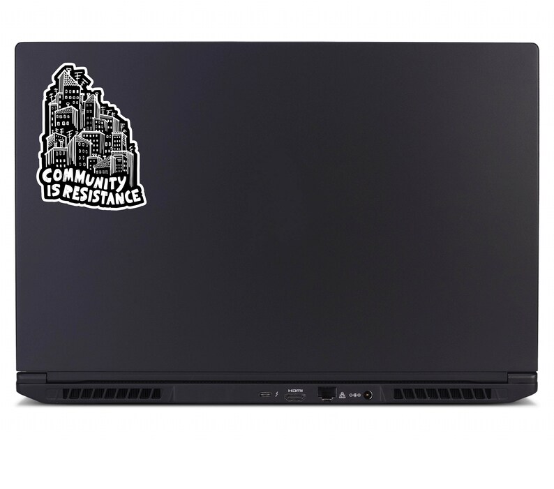 May include: Black laptop with a sticker featuring a cityscape design in white and black. The sticker reads "COMMUNITY IS RESISTANCE" in bold, capital letters. The laptop is closed, showing its back, with a sleek, matte finish.