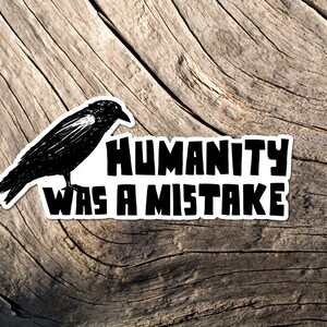 May include: A black and white sticker featuring a crow illustration and the text "HUMANITY WAS A MISTAKE." The sticker is placed on a weathered, light brown wooden surface.