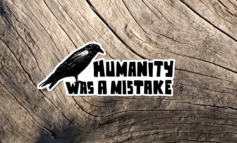 May include: A black and white sticker featuring a crow illustration and the text "HUMANITY WAS A MISTAKE." The sticker is placed on a weathered, light brown wooden surface.