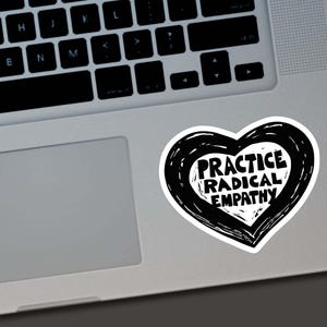 May include: A white sticker in the shape of a heart with a black outline. The sticker has the words "PRACTICE RADICAL EMPATHY" in a bold, black font. The sticker is placed on a silver laptop.