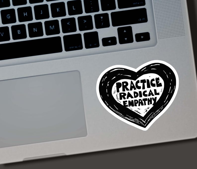 May include: A white sticker in the shape of a heart with a black outline. The sticker has the words "PRACTICE RADICAL EMPATHY" in a bold, black font. The sticker is placed on a silver laptop.