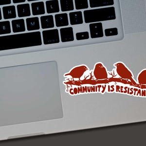May include: A sticker featuring four brown birds perched on a branch. The text "COMMUNITY IS RESISTANCE" is below the birds. The sticker is on a laptop.