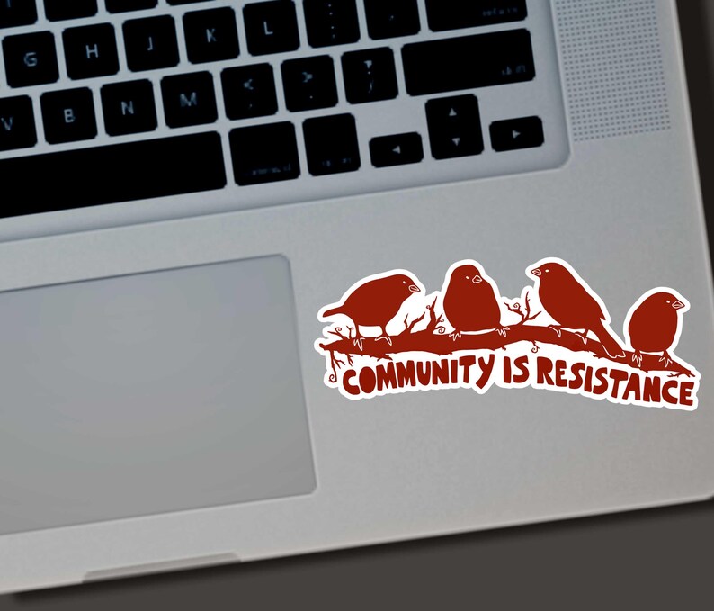 May include: A sticker featuring four brown birds perched on a branch. The text "COMMUNITY IS RESISTANCE" is below the birds. The sticker is on a laptop.