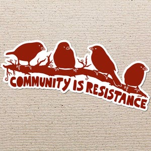May include: A sticker featuring four red birds perched on a brown branch. The text "COMMUNITY IS RESISTANCE" is written in white below the birds. The sticker has a white border and is set against a textured beige background.